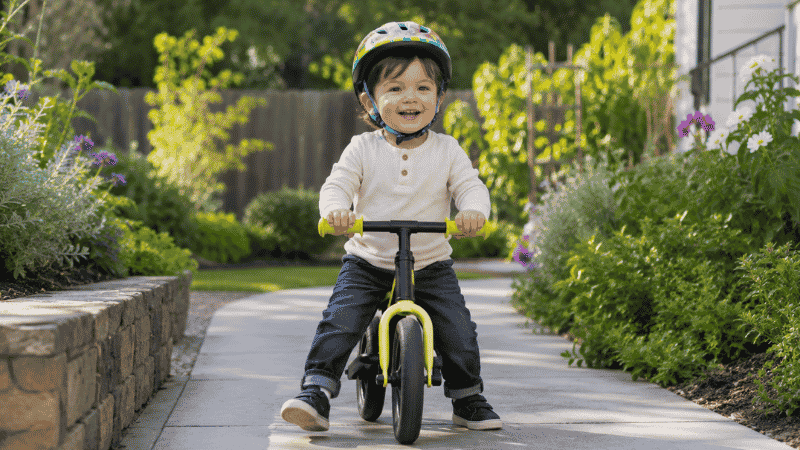 A 2-year-old toddler riding a balance bike outdoors on a sunny day wearing a safety helmet