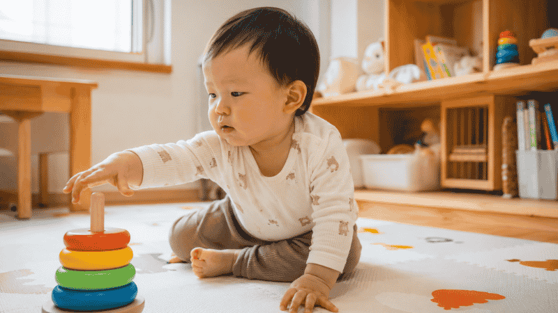 A one-year-old toddler reaching for colorful wooden stacking rings on a soft play mat in a bright living room