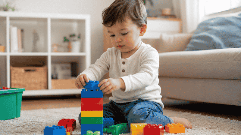 A 2-year-old toddler sitting on a rug stacking colorful DUPLO blocks with focused concentration in a bright living room