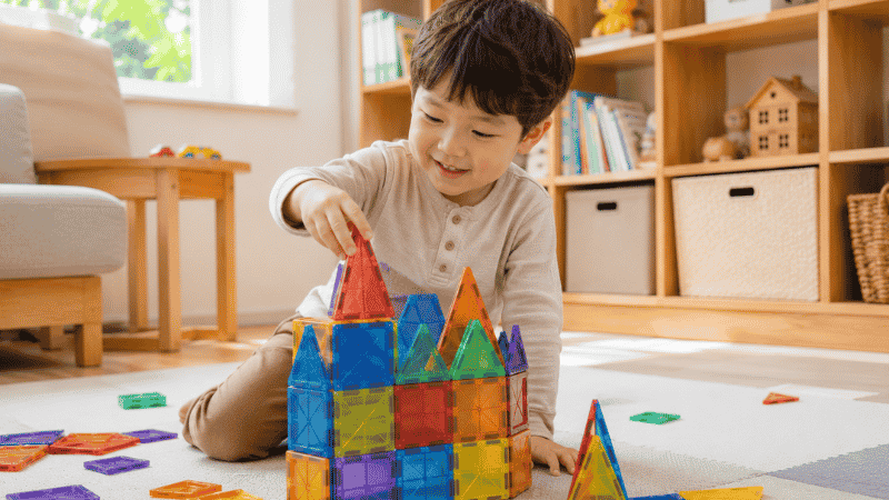 A 3-year-old child building a colorful structure with magnetic tiles on a play mat in a bright living room