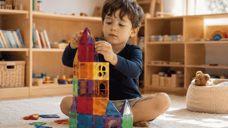 A 4-year-old child building a tall colorful structure with magnetic tiles on a soft rug in a bright playroom