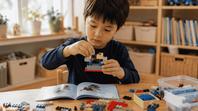 A 5-year-old child carefully building a colorful LEGO set at a small wooden table in a bright playroom