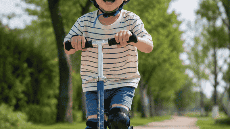 A confident 4-year-old child riding a two-wheel scooter outdoors on a sunny path wearing a helmet and knee pads