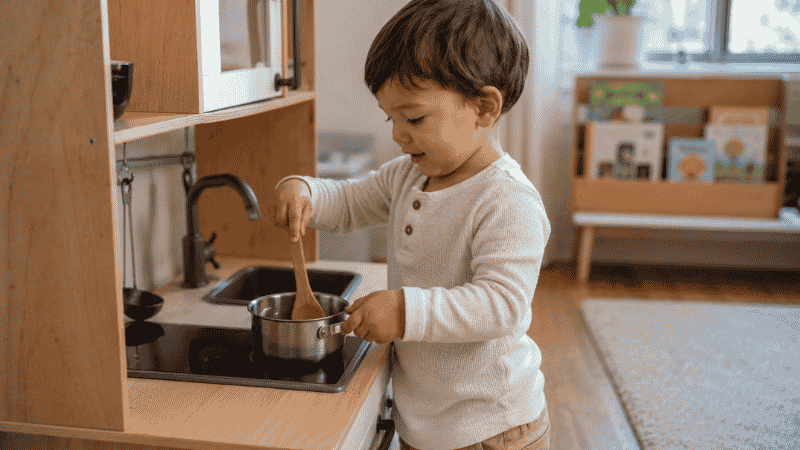A 2-year-old toddler pretending to cook at a small wooden play kitchen with a wooden spoon