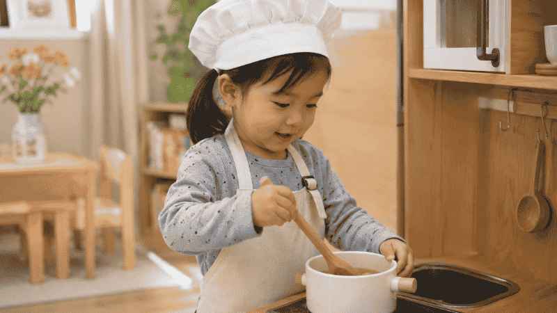A 3-year-old girl playing with a wooden toy kitchen, wearing a chef hat and stirring a pot
