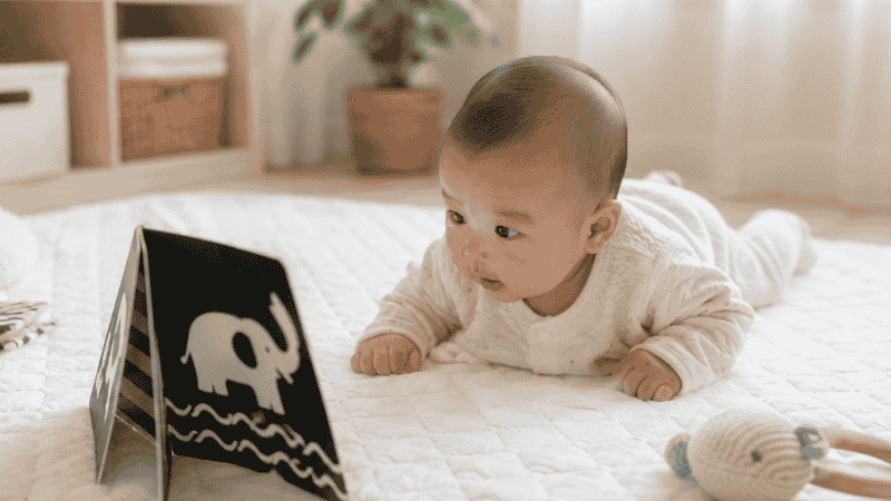A 4-month-old baby doing tummy time on a white play mat, looking at a high-contrast black and white book for sensory development