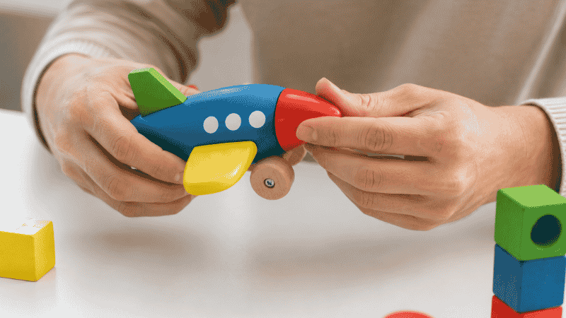 A parent's hands carefully inspecting a colorful wooden toddler toy on a white table, checking for loose parts and sharp edges. Clean minimal background, soft natural light. Photorealistic, close-up angle showing attentive care.