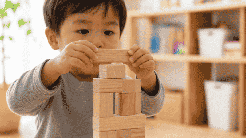 Young child placing a wooden unit block on a tower, building with classic wooden blocks at age 3