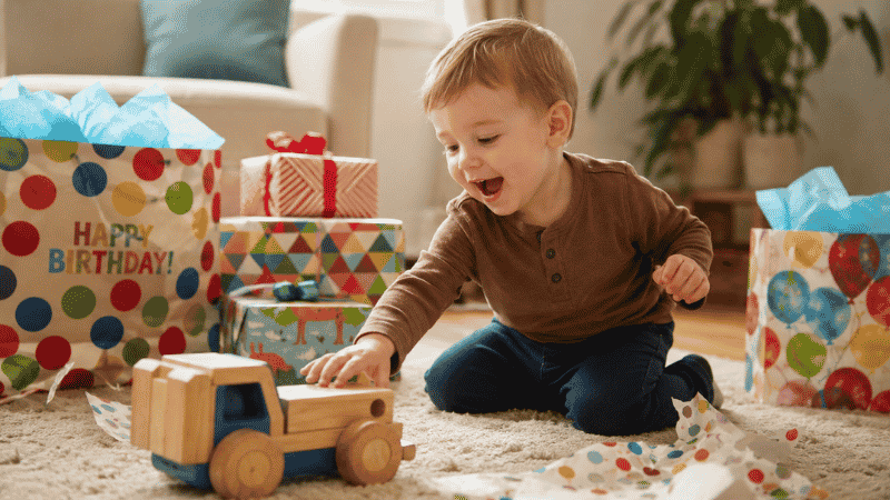 A happy 2-year-old toddler sitting on a living room rug reaching excitedly toward colorful birthday gifts on a warm birthday morning
