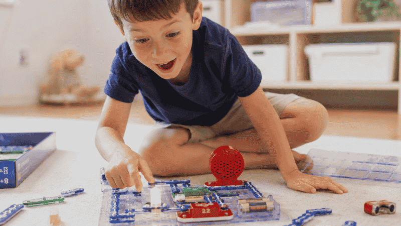 A 5-year-old child connecting snap circuit pieces on the floor, making a light bulb glow as part of a STEM learning activity