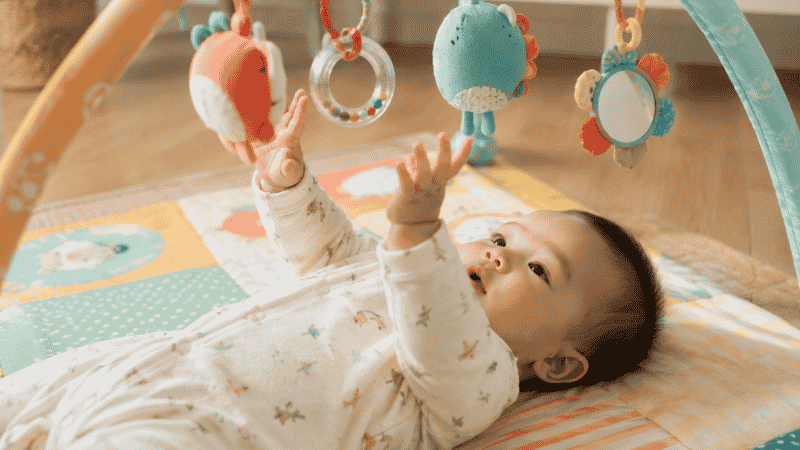 A 6-month-old baby lying on a colorful activity mat reaching up toward hanging toys on a play gym in a bright home setting