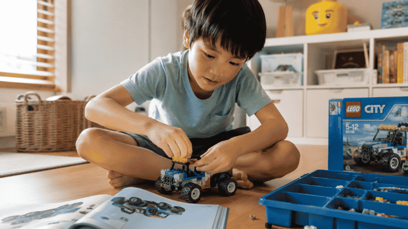 A 6-year-old child sitting on a wooden floor carefully building a colorful LEGO set following an instruction booklet in a bright playroom