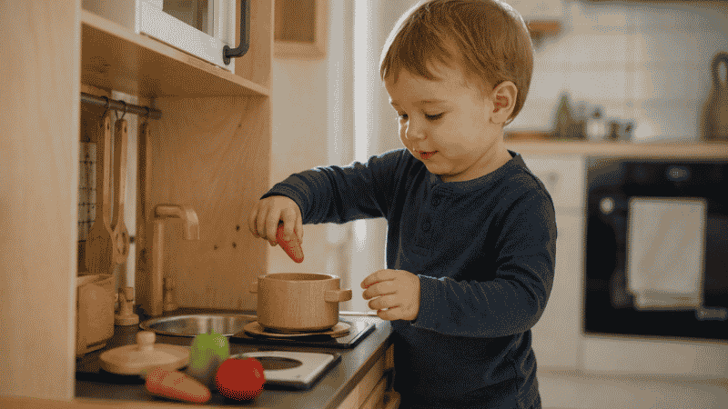 A 2-year-old toddler carefully placing a wooden toy vegetable into a pot at a small wooden play kitchen with focused concentration