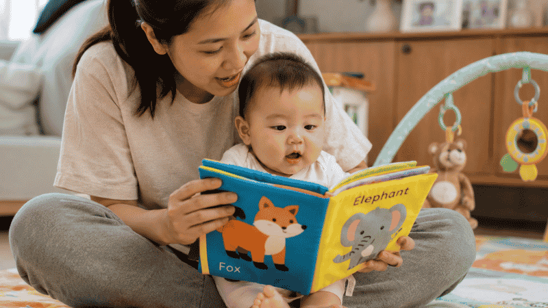 A mother holding a colorful soft fabric book in front of her 6-month-old baby on her lap, reading and narrating together for language and bonding development