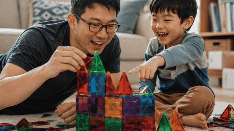 A father and 6-year-old child laughing together while building a colorful structure with magnetic tiles in a warm living room