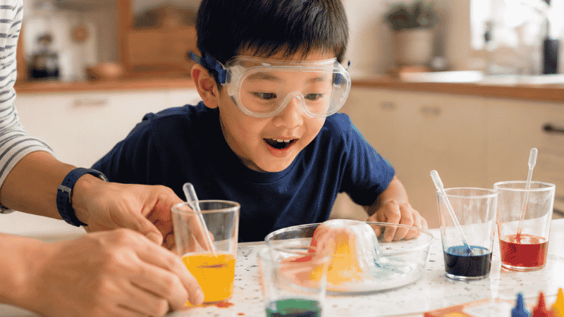 A 6-year-old child wearing safety goggles excitedly conducting a simple science experiment at a kitchen table with a parent nearby
