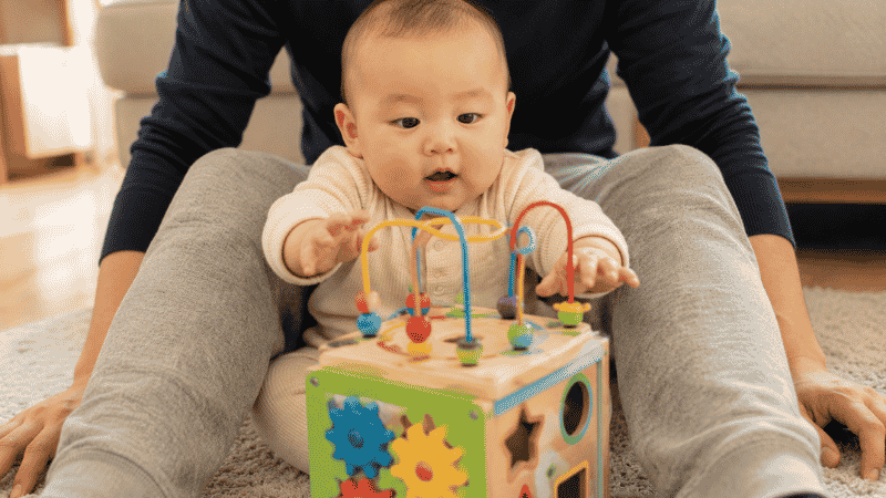 A 6-month-old baby sitting supported between a parent's legs, reaching toward a colorful activity cube for sitting play and fine motor development