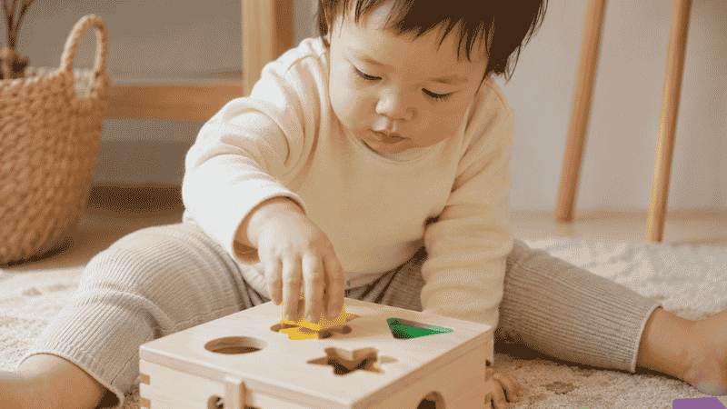 A 2-year-old toddler pushing a colorful wooden shape into a shape sorter, developing early STEM problem-solving skills