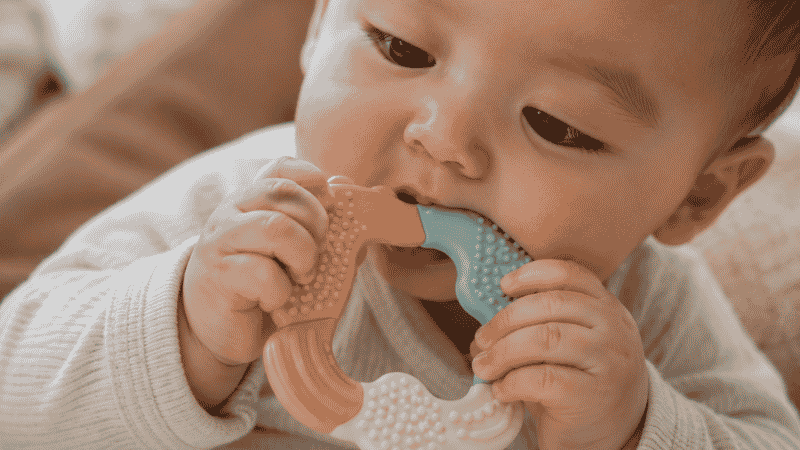 A 6-month-old baby holding and exploring a colorful food-grade silicone teether with textured surfaces for teething relief and sensory development