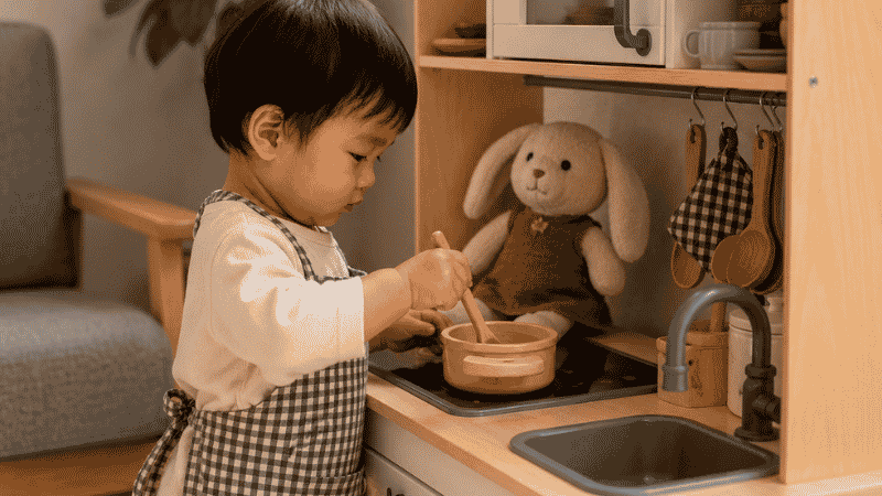 A 2-year-old toddler stirring a wooden spoon in a small pot at a wooden play kitchen with a stuffed rabbit watching nearby