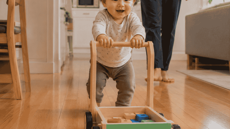 An 18-month-old toddler pushing a sturdy wooden walker wagon across a hardwood floor taking confident early steps with a parent nearby