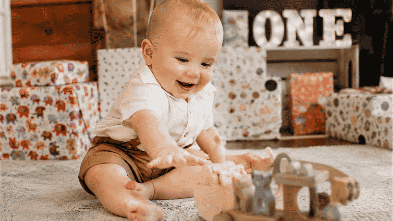 A 1-year-old baby sitting on a soft rug reaching curiously toward a wooden toy surrounded by colorful birthday gifts with a big smile