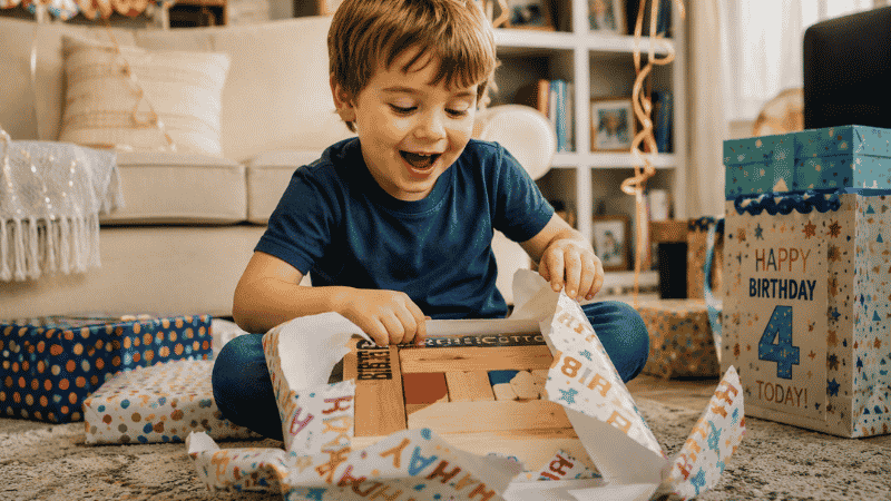 A happy 4-year-old child sitting on a rug tearing open colorful birthday wrapping paper to reveal a wooden toy with excitement