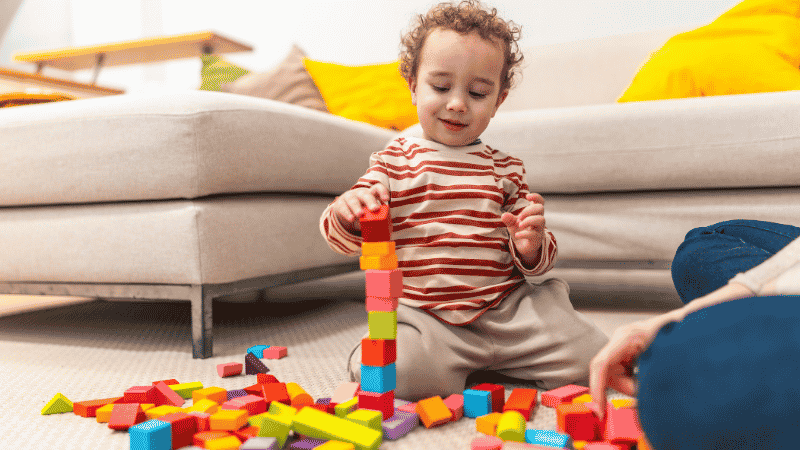 A focused 2-year-old toddler sitting on a living room rug building a colorful tower using magnetic tiles, demonstrating fine motor skills.