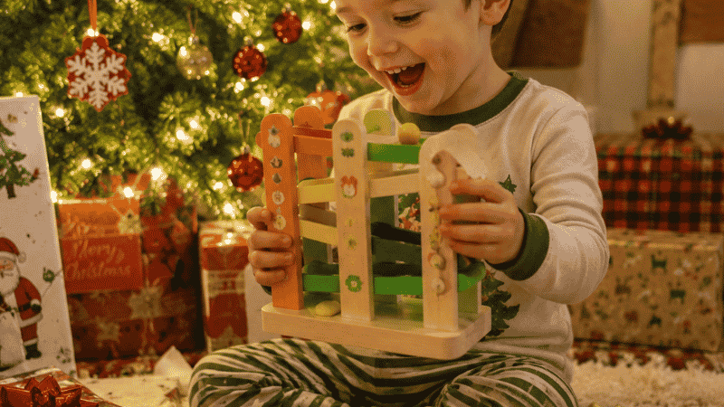 A 3-year-old child sitting next to a warmly lit Christmas tree holding a colorful wooden toy with a delighted expression surrounded by wrapped gifts