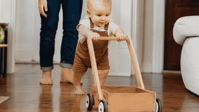 A 1-year-old baby taking confident early steps while pushing a sturdy wooden walker wagon across a hardwood floor with a parent nearby