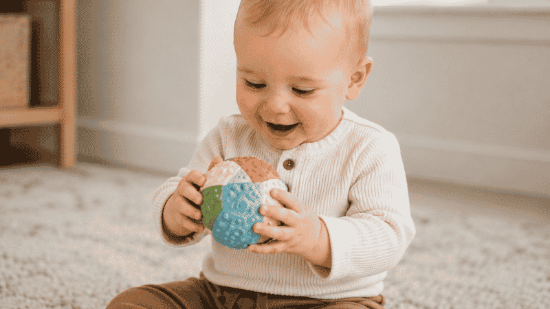 A 1-year-old baby sitting on a soft rug holding a colorful textured sensory ball in both hands with a curious and delighted expression
