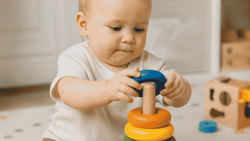 Close-up of a 1-year-old baby's hands carefully placing a colorful ring onto a stacking toy on a soft play mat with concentrated expression