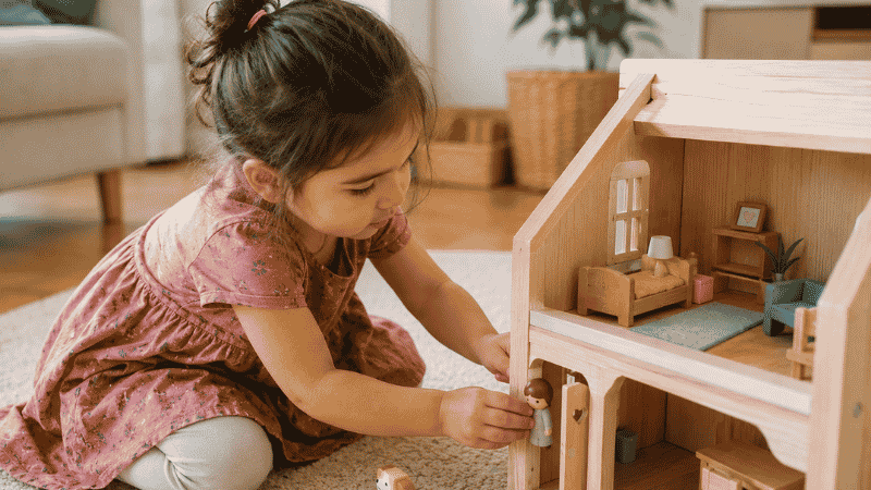 A 3-year-old girl carefully placing a small wooden figure into a wooden dollhouse with focused concentration in a cozy home setting