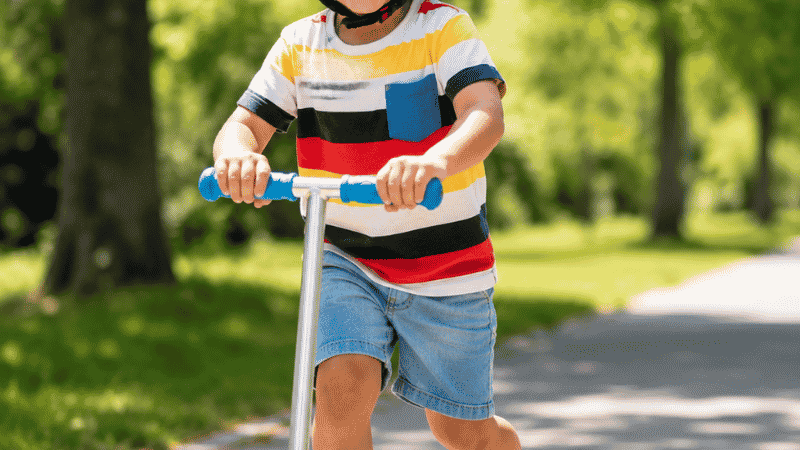 A confident 3-year-old child riding a three-wheel scooter on a sunny outdoor path wearing a bright helmet with a big smile