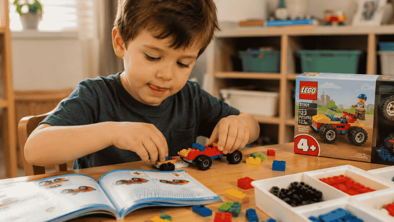A 4-year-old boy sitting at a small table carefully building a colorful LEGO set following an instruction booklet with concentrated expression