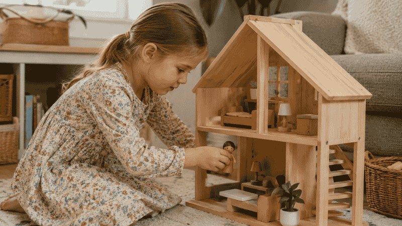 A 4-year-old girl kneeling in front of a wooden dollhouse carefully moving a small figure through a room with an imaginative focused expression