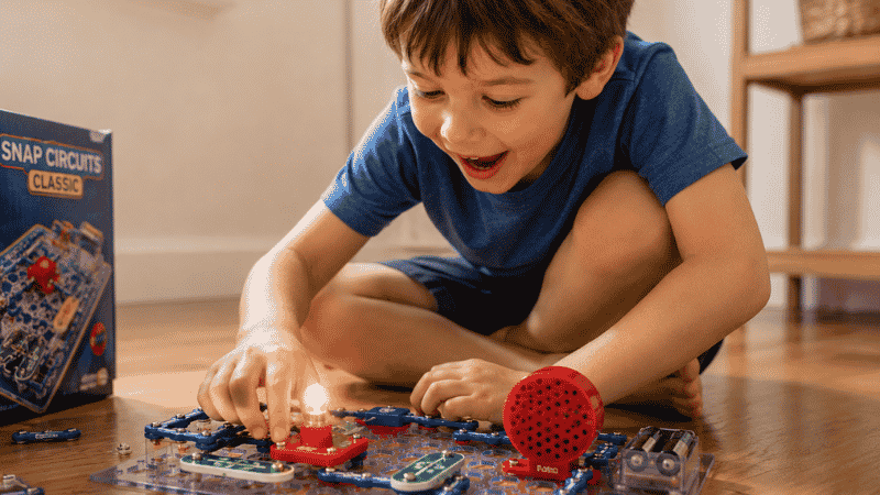 A 5-year-old boy connecting snap circuit pieces on the floor looking amazed as a small light bulb glows from his completed circuit