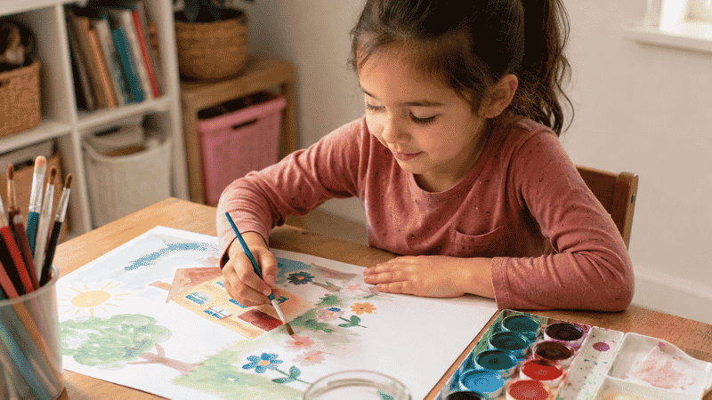 A 5-year-old girl carefully painting with a watercolor set at a small wooden table with natural light, looking focused and proud of her artwork