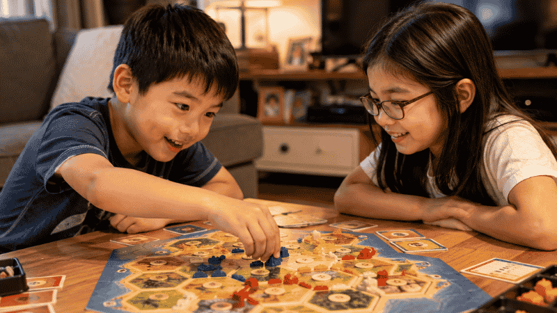 A 5-year-old child and older sibling leaning forward with competitive focus while playing a colorful strategy board game at a low table