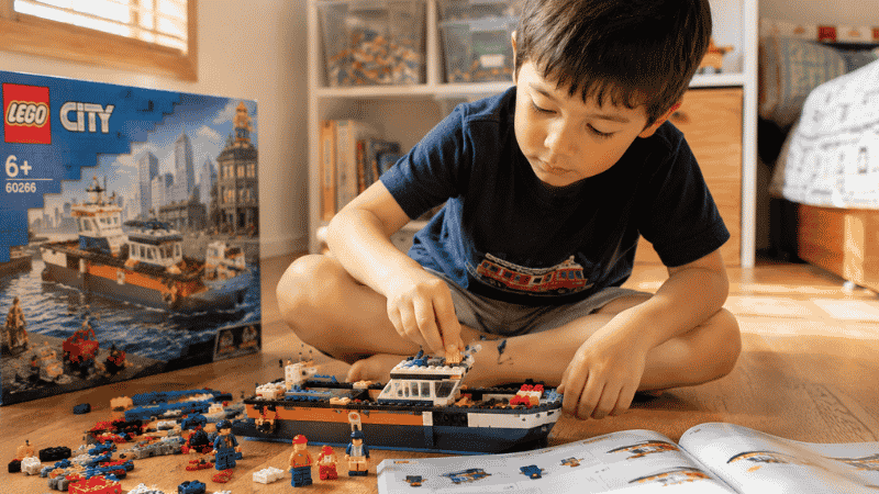 A 6-year-old boy sitting on a wooden floor deeply focused on building a detailed LEGO City set following an instruction booklet beside him