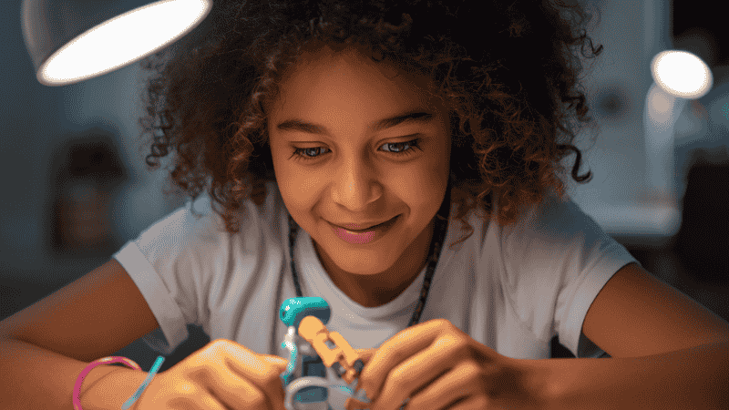 A focused 10-year-old girl sitting at a desk, using a small screwdriver to assemble a DIY robotic arm kit with wires and mechanical parts