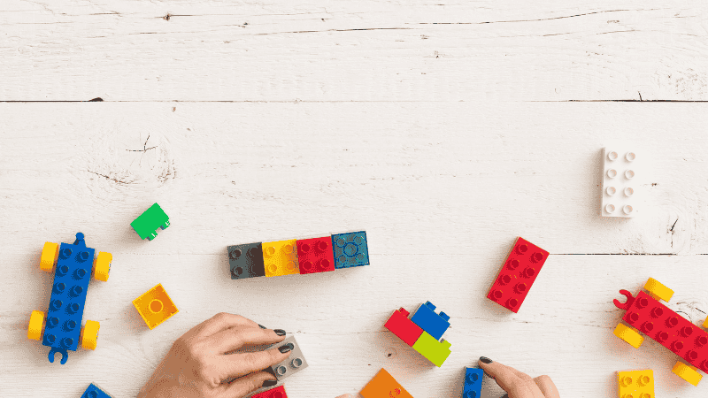 A parent's hand gently pointing to a step in a LEGO 4 plus instruction manual while a preschooler holds a bright red brick, ready to snap it into place.