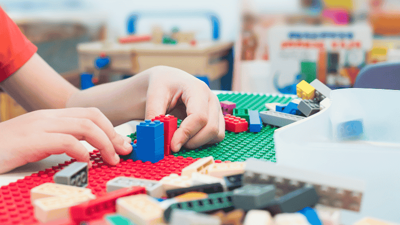 A 4-year-old child sitting on a colorful play rug, smiling and proudly holding up a partially finished LEGO vehicle they built themselves.