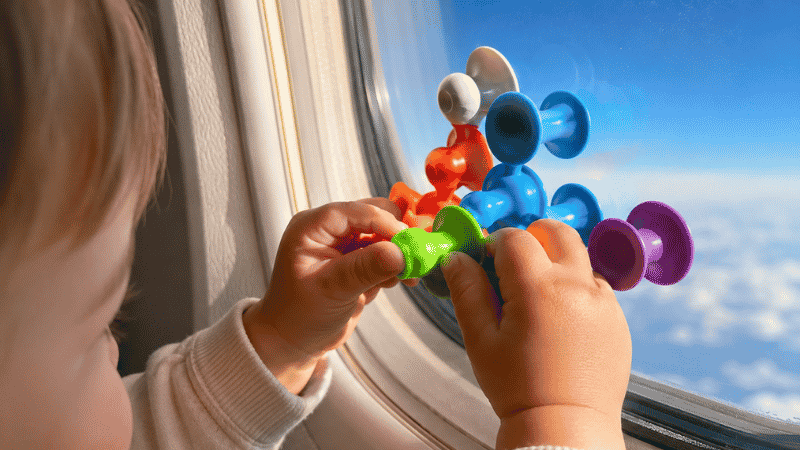 Close-up of a toddler's small hands pressing a colorful suction cup toy attached to an airplane window with blue sky visible behind