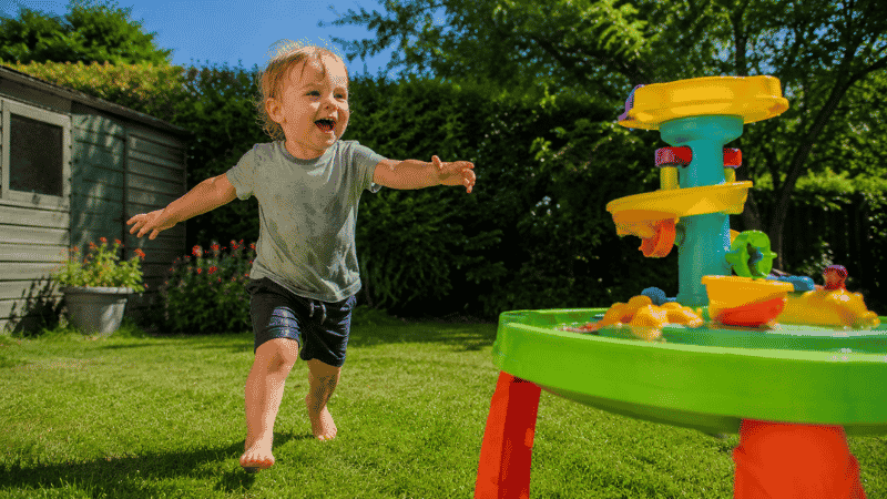 A happy 2-year-old toddler running barefoot across a green lawn toward a colorful sand and water table on a sunny afternoon