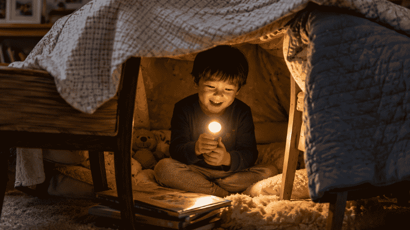 A 4-year-old child sitting inside a cozy blanket fort holding a flashlight with warm golden light illuminating their delighted face