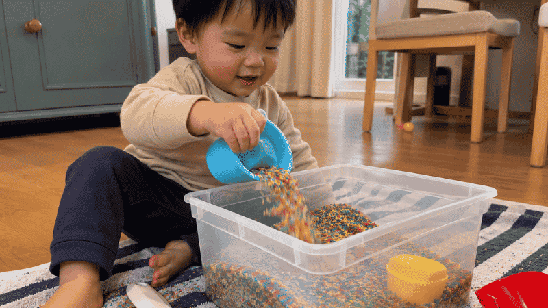 A 2-year-old toddler sitting beside a clear plastic sensory bin filled with colorful dried rice scooping and pouring with a small cup