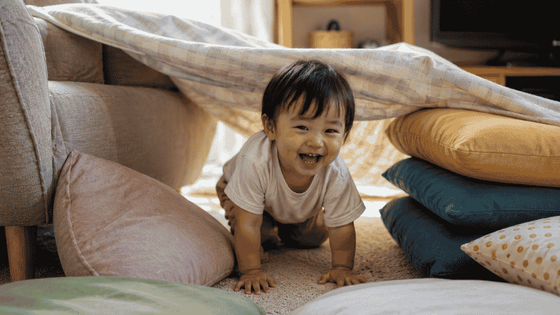 A laughing 2-year-old toddler crawling through a blanket tunnel surrounded by couch cushions as a living room obstacle course on a rainy day
