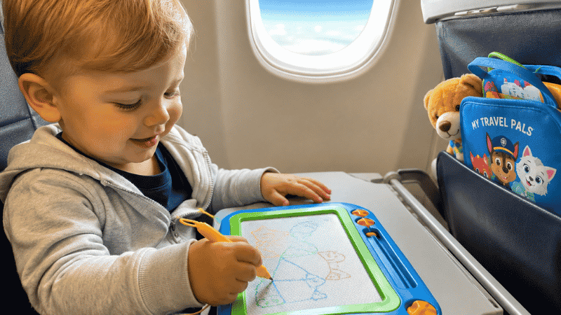 A happy 2-year-old toddler sitting in an airplane seat focused on drawing on a colorful magnetic drawing board on the tray table