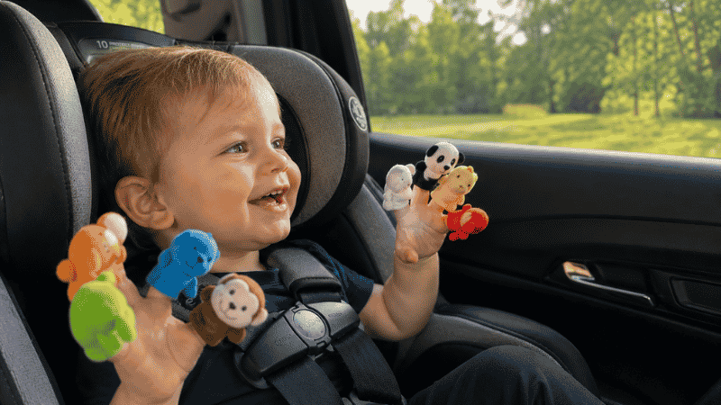 A cheerful 2-year-old toddler sitting in a car seat happily playing with colorful finger puppets on both hands while looking out the window